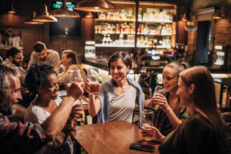 Group of young adults sit around a table, each holding a glass of beer with their drinks raised.