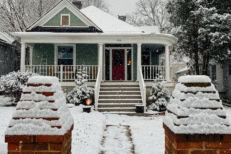 Snow-covered single-family home with a front porch and red door during winter snowfall in a residential neighborhood.