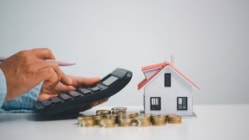 Hands using a calculator next to a small house model and stacked coins on a table, illustrating mortgage rate calculations, home loan payments, housing affordability, and real estate financing costs.