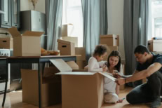 Family unpacking moving boxes in a new home with parents and child sitting on the floor, illustrating homeownership, moving day, real estate purchase, and settling into a new house.