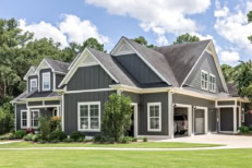 A large gray craftsman new construction house with a landscaped yard and leading pathway sidewalk on a sunny day with blue skies and clouds.