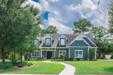 A large gray craftsman new construction house with a landscaped yard and leading pathway sidewalk on a sunny day with blue skies and clouds.