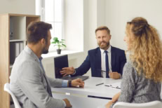 Young family couple sitting at desk with real estate agent, looking at laptop screen, choosing new house. Husband and wife exploring real estate options for purchase or rent with professional real estate agent.