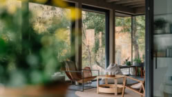 Photo of modern wooden cabin interior featuring a minimalist dining area bathed in natural light. Large glass windows provide a serene view of the forest with vibrant autumn foliage, creating a peaceful nature-inspired atmosphere. The clean design includes wooden furniture, a spacious table, and contemporary chairs, offering a tranquil and cozy retreat perfect for relaxation or a getaway. Ideal for concepts of modern living, eco-friendly homes, sustainable architecture, and indoor-outdoor harmony